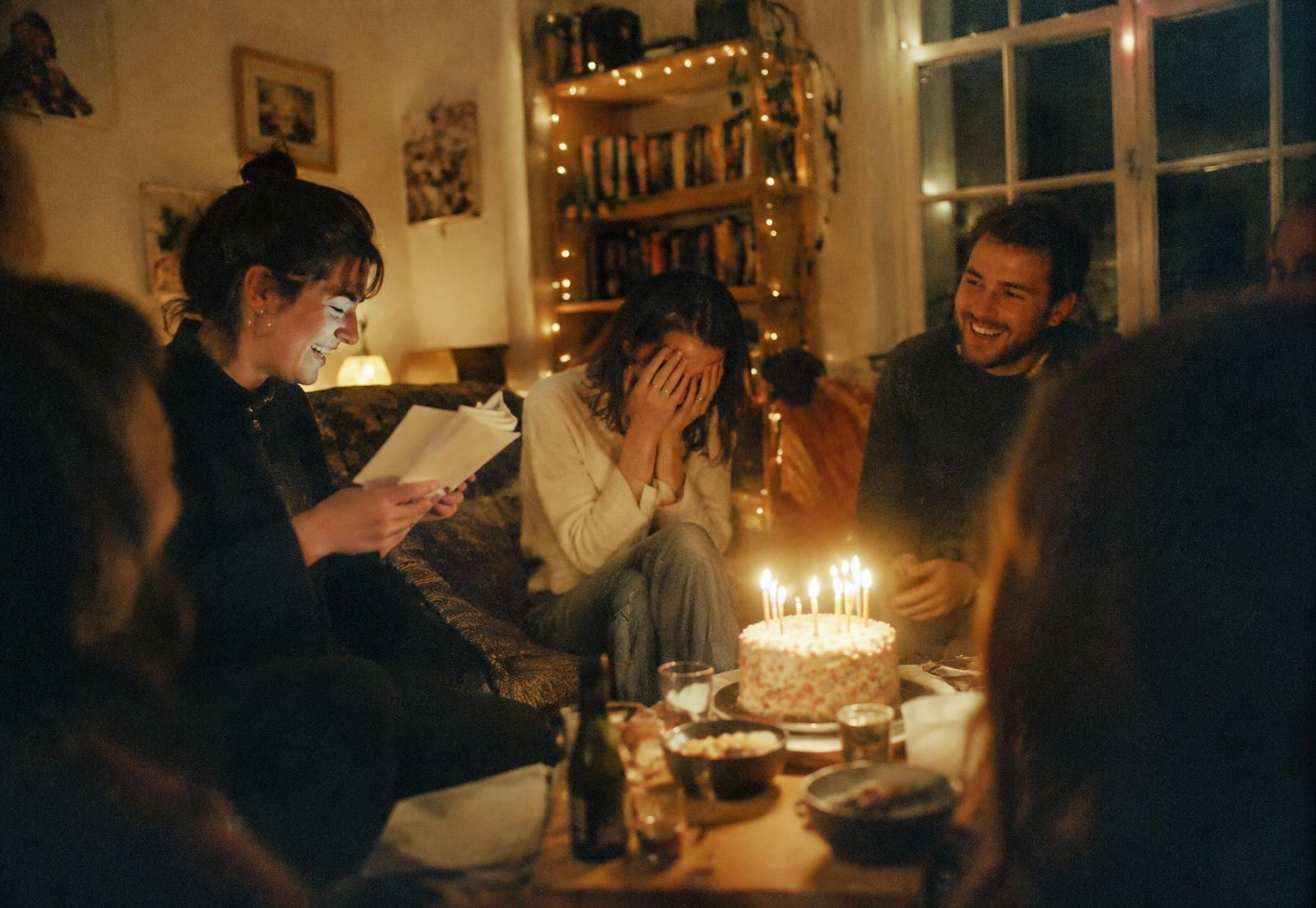 Friends in a living room laughing while the birthday person hides their face